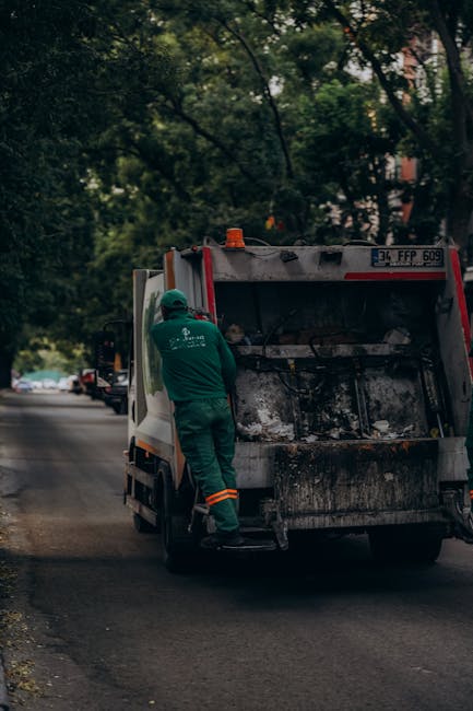 A waste collection worker dressed in a dark green uniform and wearing a protective helmet is standing beside the open rear compartment of a small rubbish collection truck parked on a city street. The 