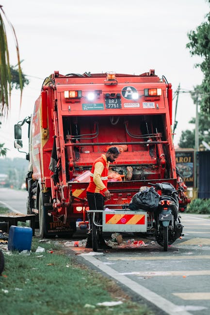 A waste collection worker dressed in a dark green uniform and wearing a protective helmet is standing beside the open rear compartment of a small rubbish collection truck parked on a city street. The 